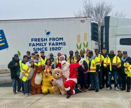 A large group of people in safety vests pose in front of a Perdue truck, joined by people in holiday costumes including Santa and characters dressed as a bear and a reindeer.
