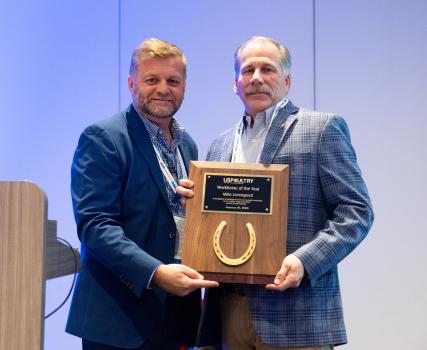 Two people standing together hold a wooden award plaque featuring a horseshoe and a title plate during an indoor recognition event.