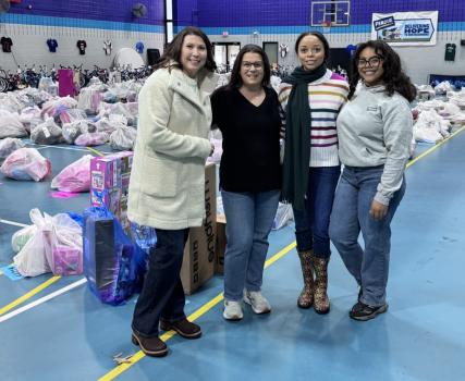Four people stand together inside a gymnasium filled with large bags of donated gifts and toys arranged in rows for a holiday giving event.