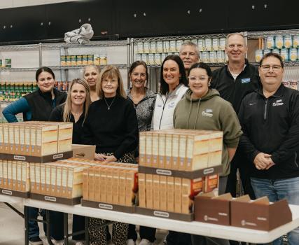 A group of people stands behind a table filled with boxed food items in a pantry setting, with shelves of canned goods lining the wall behind them.