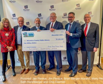 A group of people stand in front of a Wicomico County Public Library backdrop holding an oversized check for one million dollars, presented to the Wicomico County Public Library by Perdue Farms.