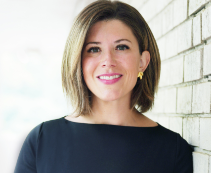 Woman in dark blue top smiling at camera against white brick background.