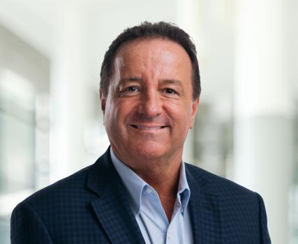 Man in dark blazer and light blue shirt standing in front of blurred interior background.