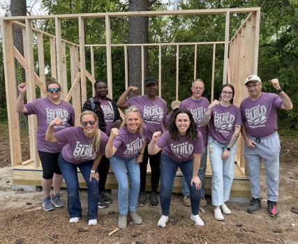 Group of people wearing purple shirts standing in front of wooden frame structure outdoors.