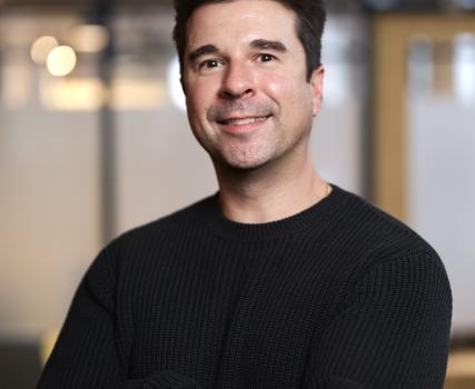 Man in black crewneck shirt standing in front of blurred interior background.