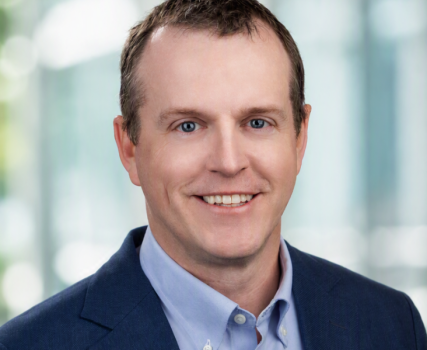 Person wearing dark blazer and light blue shirt in front of blurred indoor background.