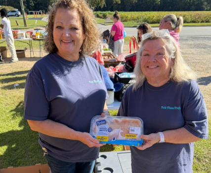 Two women in dark polo shirts holding blue award at outdoor event with trees and people in background.
