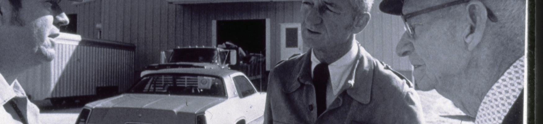 Three people stand and talk outside a Perdue facility, with a car parked in front of the building and large industrial structures in the background.