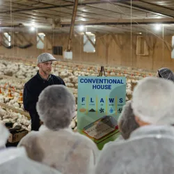 Person in black jacket speaking to an audience in an industrial facility with stacked boxes in background.