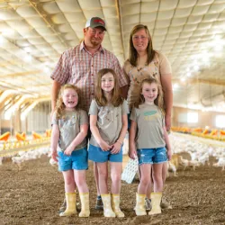 Family of five standing together in a poultry barn with chicken houses visible in background.