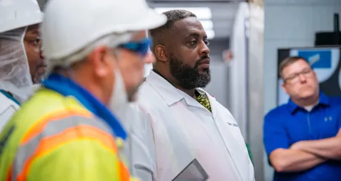 Several people wearing safety gear and work attire stand together inside an industrial facility, appearing to listen during a walkthrough or briefing