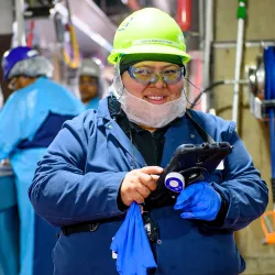 Worker in blue jacket and safety gear holding equipment in industrial facility.