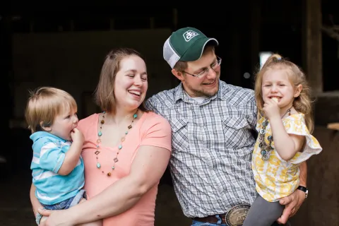 Family of four posing together indoors, two adults holding two toddlers in casual clothing.