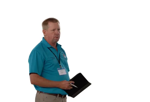 Person wearing teal polo shirt holding a tablet, standing against white background.