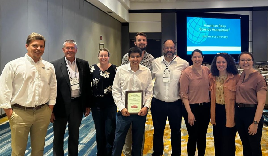 Group of eight people standing together in an indoor facility with a presentation screen in the background, one person holding a framed award or certificate.