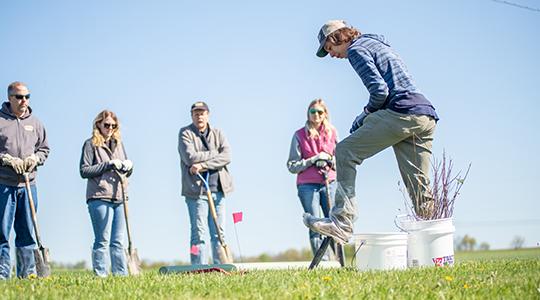 Group of people standing outdoors watching person jump in grassy field under clear sky.