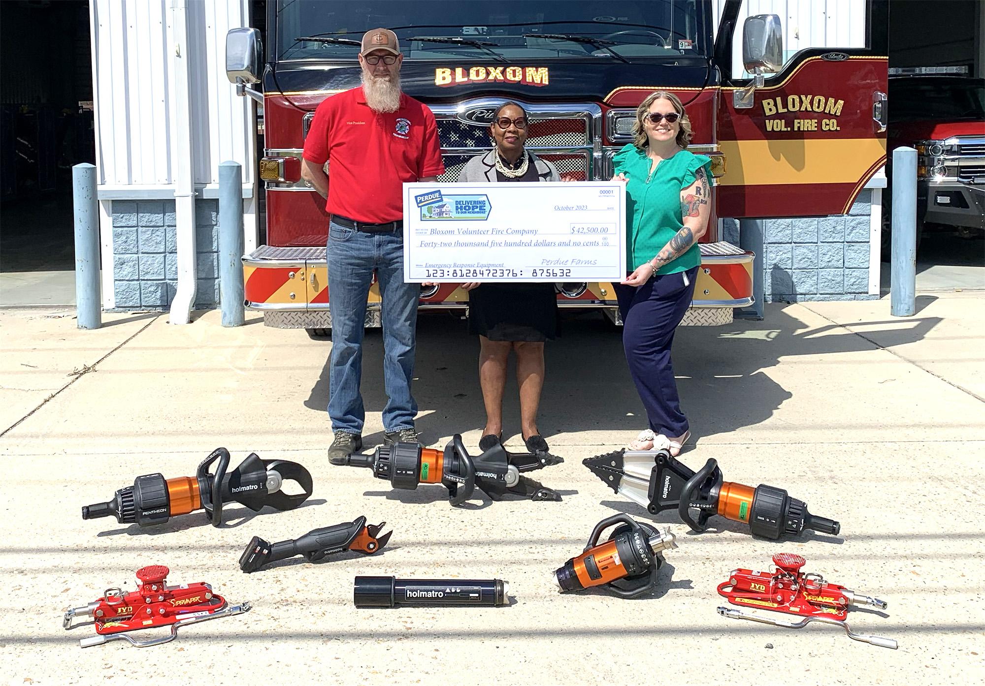 Two people in red and blue shirts standing behind a large check, with tools and equipment displayed on the ground in front of them.