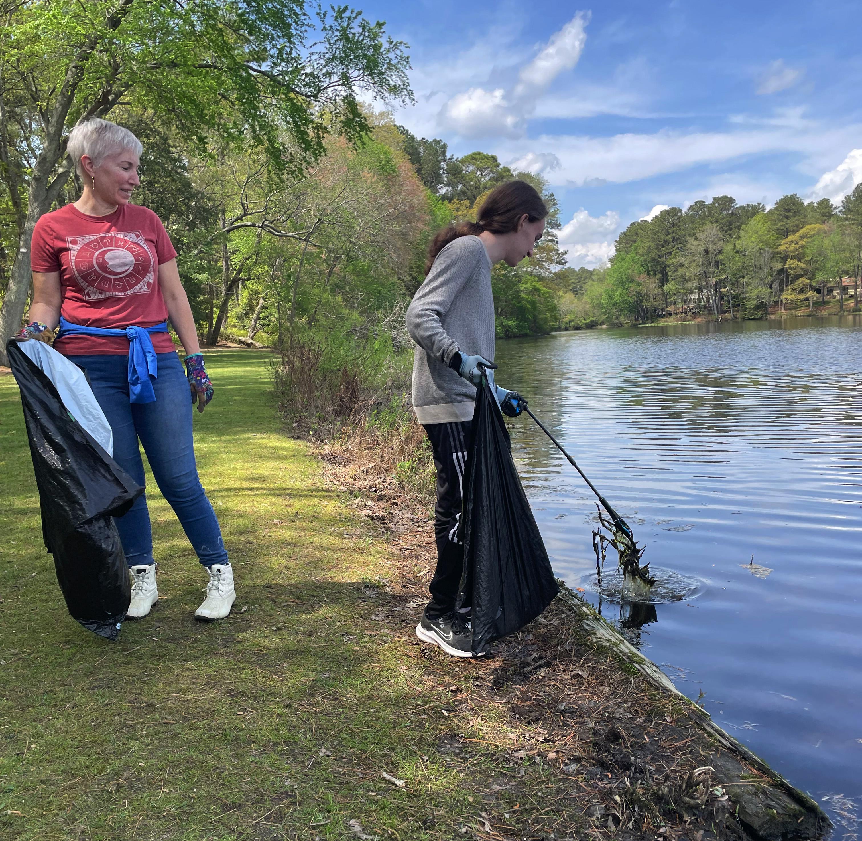 Two people fishing by a calm lake on a sunny day, one wearing a pink hat and red jacket, the other in gray and black clothing.