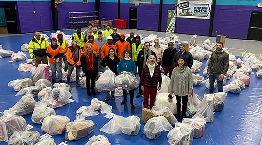 Group of people standing in circle surrounded by white bags on blue floor in warehouse setting