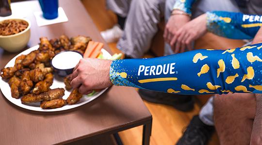 Person holding Perdue branded item next to plate of cooked chicken wings and dipping sauce.