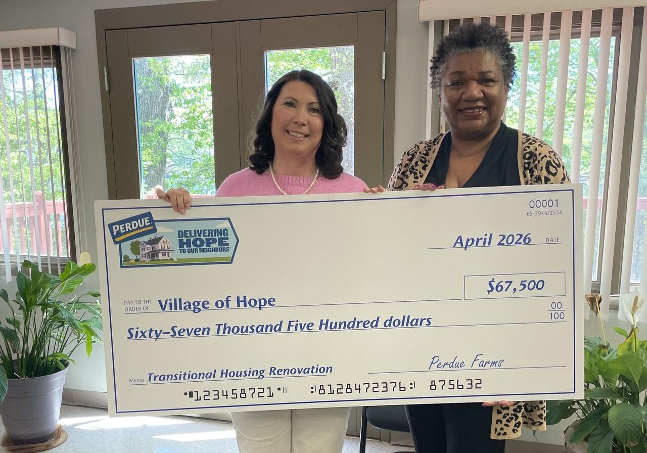 Two people holding a large check for Village of Ilion Community School District in a home interior.