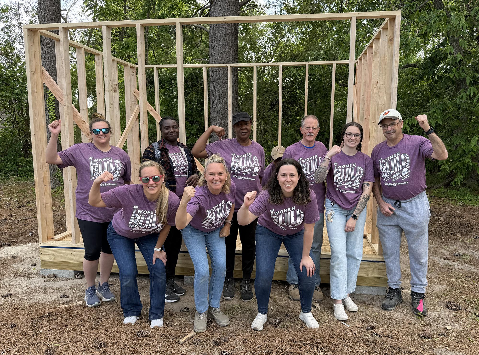 Group of people wearing purple shirts standing in front of wooden frame structure outdoors.