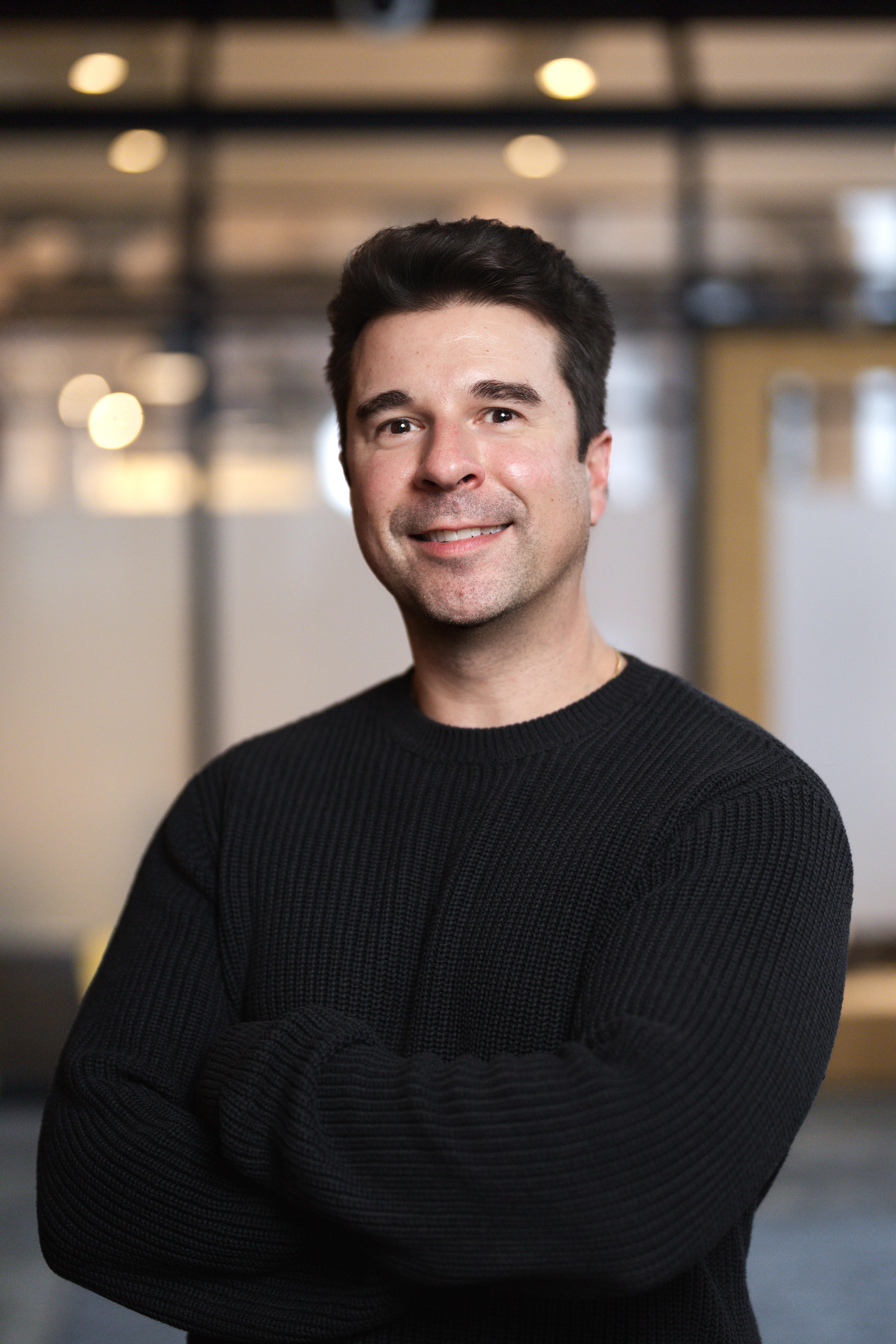 Man in black crewneck shirt standing in front of blurred interior background.