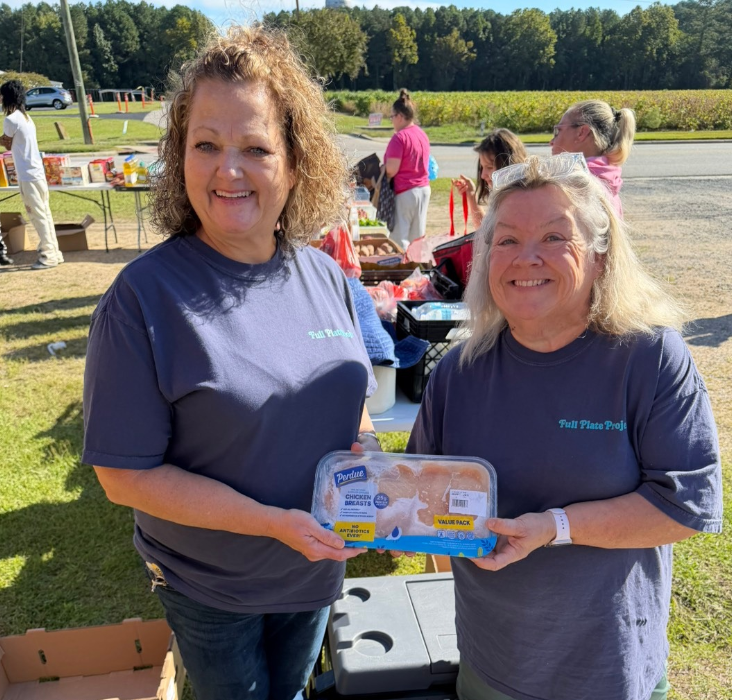 Two women in dark polo shirts holding blue award at outdoor event with trees and people in background.