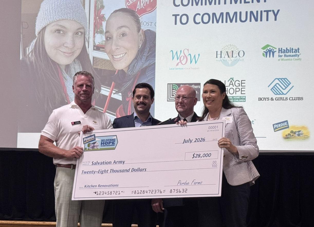Four people in white Perdue branded shirts holding oversized check at community event.
